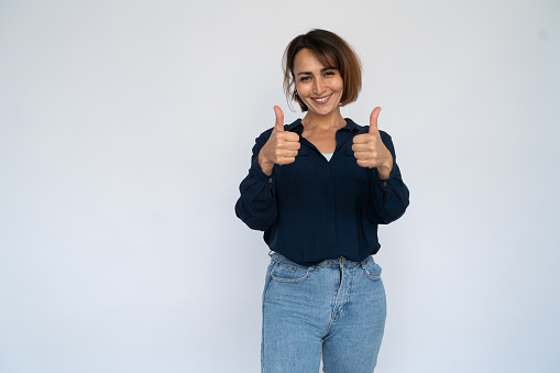 Excited woman making like gesture. Female model in dark blue shirt showing thumbs up with both hands. Portrait, studio shot, advertising concept