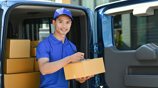 Smiling delivery man sitting in open delivery van and checking checking address details of client on clipboard.