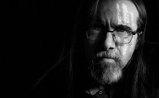 Mid adult man with beard, long hair, white war paint on his face and confidence in his eyes. Black and white portrait. Dark background. Selective focus
