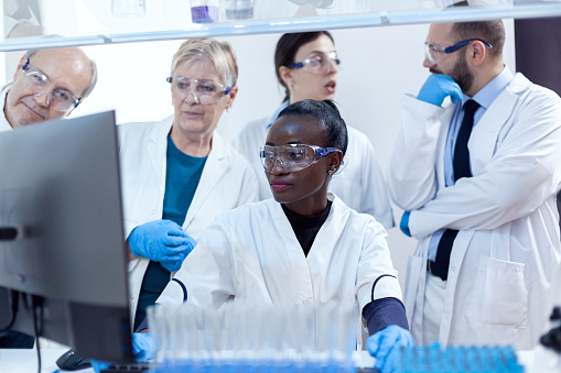 Team of scientists with multiethnicity discussing chemical formula in front of computer. Black healthcare researcher in biochemistry laboratory wearing sterile equipment.