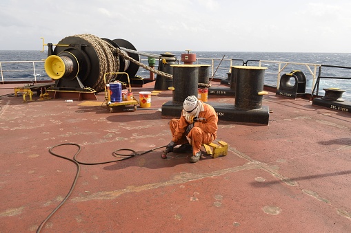 at sea, on board ship - december 21, 2022 : crew member of commercial cargo ship performing deck maintenance work while chipping  and buffing at poop deck of ship using a grinder buffing machine