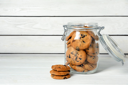 Glass jar with chocolate chip cookies on white wooden table, space for text