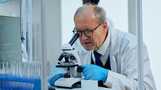 Laboratory scientist conducting experiment looking at microscope in busy modern lab. Eldery man working with various bacteria tissue, blood samples, concept of pharmaceutical research for antibiotics