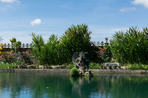 Stone monuments, Indonesian culture and history - Tirta Gangga park with lush green tropical trees, pond, fountain, foothpaths and statues