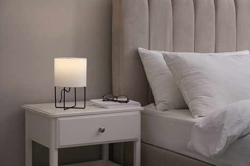 Stylish lamp, notebooks and glasses on bedside table indoors. Bedroom interior elements