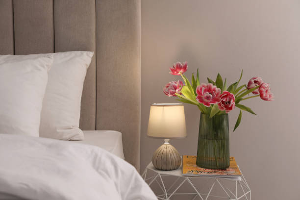 stylowa lampa, kwiaty i czasopismo na stoliku nocnym w pomieszczeniu. elementy wnętrza sypialni - cozy bedside zdjęcia i obrazy z banku zdjęć