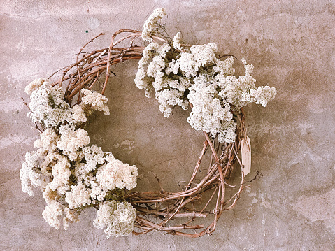 Horizontal closeup photo of a floral wreath made from woven vines and two bunches of dried white flowers, hanging on a rustic pink and grey rendered wall inside a Florist shop in Summer. Hunter Valley, NSW