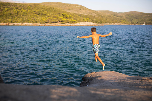 A happy young boy is in mid-air jumping off a cliff into the sea