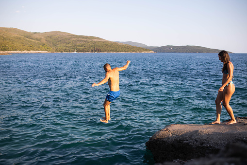 A playful young couple is jumping off a cliff into the sea