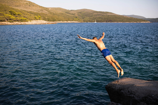 A young man is in mid-air jumping off a cliff into the sea