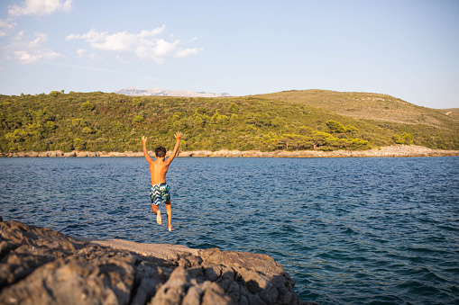 A happy boy is in mid-air jumping off a cliff into the sea with his arms up in the air