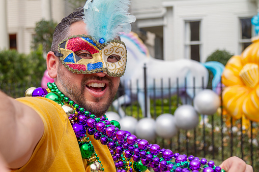 A young latin tourist male, wearing mask, costumes and necklaces celebrating Mardi Gras through the streets while taking a selfie with his cellphone in New Orleans. This is the most important celebration for the city.