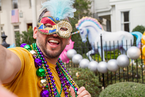 A young latin tourist male, wearing mask, costumes and necklaces celebrating Mardi Gras through the streets while taking a selfie with his cellphone in New Orleans. This is the most important celebration for the city.