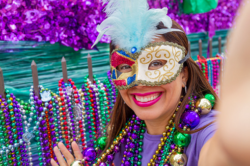 A young latin tourist female, wearing mask, costumes and necklaces celebrating Mardi Gras through the streets while taking a selfie with her cellphone in New Orleans. This is the most important celebration for the city.