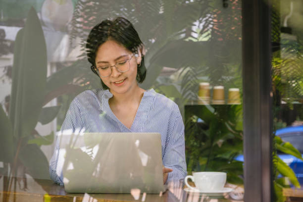 istock Young Asian women using laptop computer while sitting at coffee shop cafe, People lifestyle relaxing, Technology and working, viewed through glass with reflections 1452873907