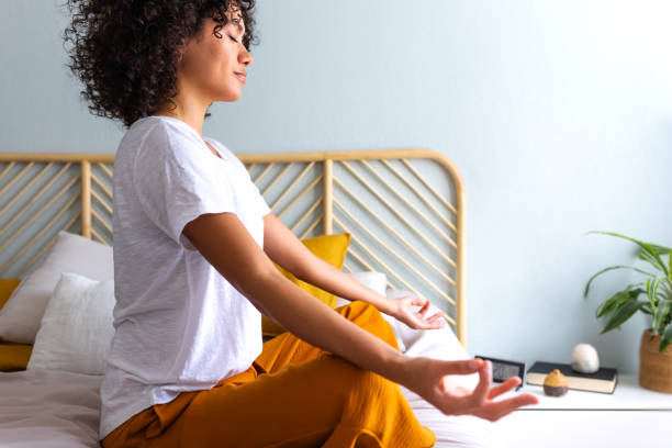 Woman meditating and breathing yoga exercises to calm mind. African American woman does meditation in bedroom. Side view of young multiracial woman relaxing on the bed. Meditating and breathing yoga exercises to calm mind. African American woman does meditation in bedroom. Wellness and meditation concept. Posture and Body Language stock pictures, royalty-free photos & images