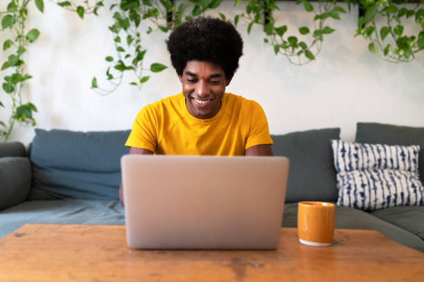 istock Smiling young african american man sitting on the sofa checking email with laptop. 1452876482