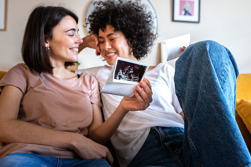 Bliss. Happy and joyful multiracial lesbian pregnant couple looking baby ultrasounds at home relaxing on the sofa. LGTBQ love and family concept.