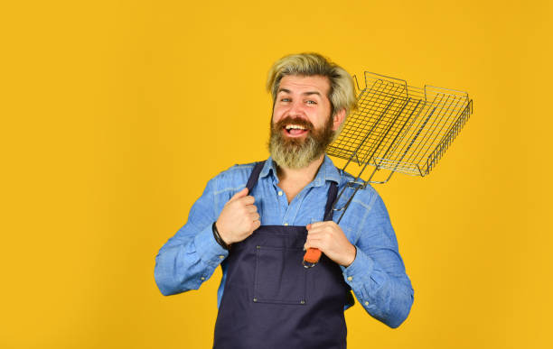 Tools for roasting meat. Man in apron hold barbecue grill. Culinary concept. Cooking utensils. Chef cooking bbq food. Summer picnic. Hipster dyed beard promoting bbq equipment. Cooking healthy