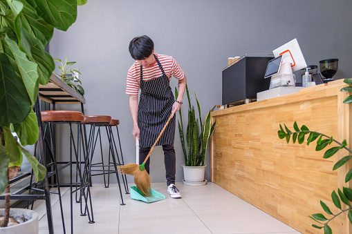 Young Asian man worker or coffee shop owner sweeping with broom and dustpan at coffee shop cafe, Small business owner