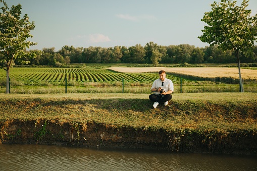 A Caucasian man sitting on a lakeshore in the countryside