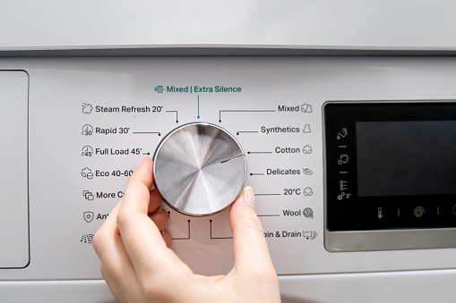 Woman hand choosing program on automatic washing machine for daily laundry at home. Turning knob on control panel for cotton washing, close up