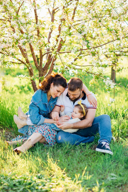 мама и папа с маленькой дочкой на лужайке в цветущем весеннем саду. - mother family vertical flower стоковые фото и изображения