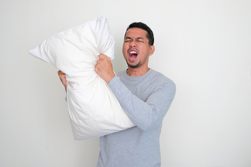 Adult Asian man screaming angry while grabbing his sleeping pillow