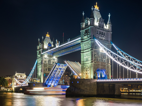 The gothic battlements of Tower Bridge illuminated at night over the River Thames as a boat travels through the open drawbridge in the heart of London, UK.