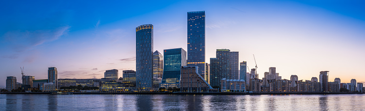 The glittering lights of Canary Wharf Docklands financial district skyscrapers and modern apartment blocks reflecting in the tranquil waters of the River Thames before sunrise in the heart of London, the UK’s vibrant capital city.