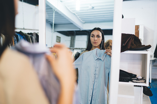 Caucasian woman with hanger trying shirt near mirror in merchandise boutique mall, attractive hipster girl posing near glass looking at own reflection while choosing brand clothing on shopping