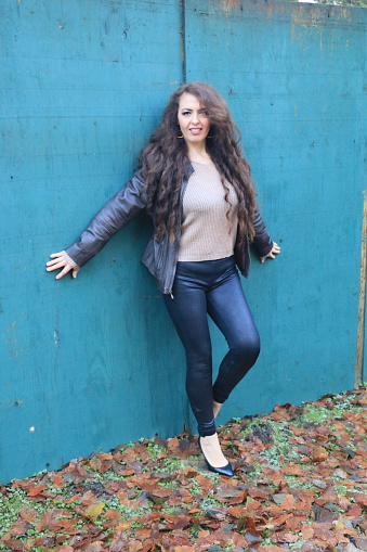 A Mexican model posing against a blue wall. She is wearing an open black jacket, beige sweater, black leather pants and shoes.