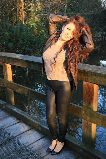 A Mexican model stretching on a wood footbridge over a pond in a public park. She is wearing an open brown leather jacket, black leather pants and beige sweater.