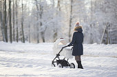 Young adult mother pushing white baby stroller and walking on snow covered sidewalk at park in cold winter day. Spending time with newborn and breathing fresh air. Enjoying peaceful stroll. Back view.