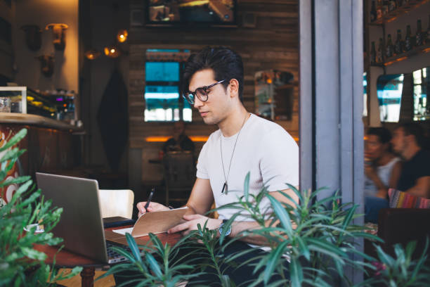 istock Serious caucasian male digital nomad writing in notepad prepare publication article for blog, clever young hipster guy student making notes learning online course via laptop computer in coffee shop 1452951443