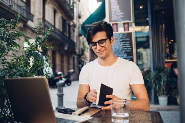 istock Handsome caucasian 20s hipster guy blogger satisfied with remote job checking notes in notepad, smiling male millennial writer in eyewear for vision correction sitting on terrace with laptop computer 1452951456