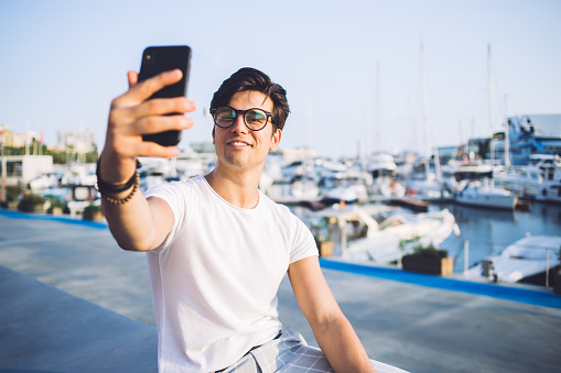 Cheerful caucasian young male generation z posing for selfie on smartphone camera sitting in marine port in city, smiling young hipster guy blogger shooting video from trip holding modern mobile phone