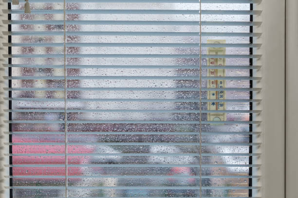 Part of the modern window with window panes covered with water drops, attached outdoor thermometer, inside view across the blinds