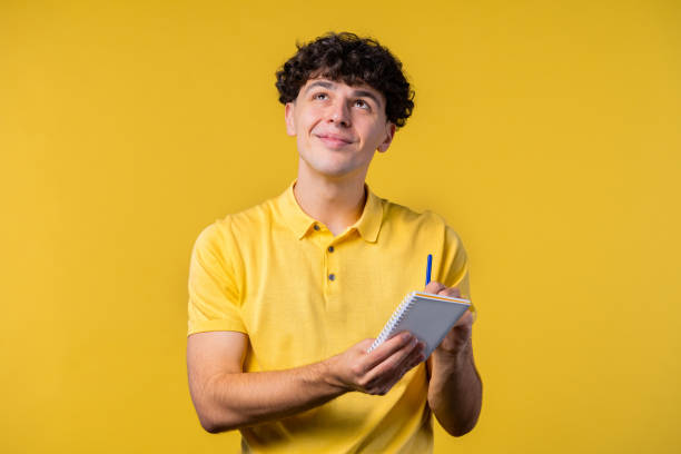 istock Curly man writing notes in planner with pen. Student guy writes future plans and to-do list in notebook for week, month, year. Keeping personal diary on yellow background. 1452970244