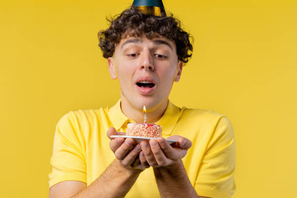 istock Happy birthday man making wish - blowing candle on cake. Curly haired guy laughs, celebrating anniversary. Young teenager on yellow background. 1452970261