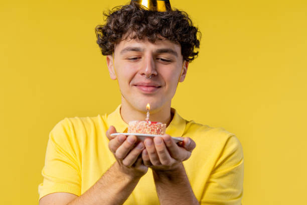 istock Happy birthday man making wish - blowing candle on cake. Curly haired guy laughs, celebrating anniversary. Young teenager on yellow background. 1452970265