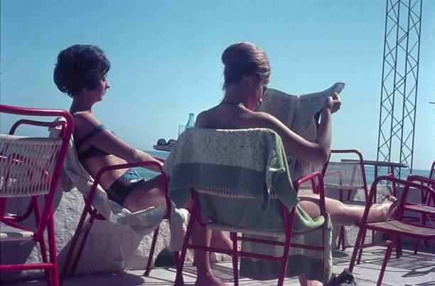 A casual moment outdoors as a woman in a hat and summer attire reads a newspaper against a bright wall under the sunlight, exuding relaxation and leisure.