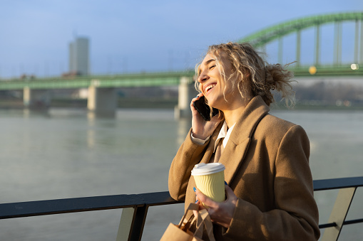 A girl with coffee and shopping bags stands by the river at the view after shopping while talking on the phone
