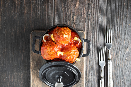 Beef meatballs with tomato sauce in back pot and forks on wooden background.