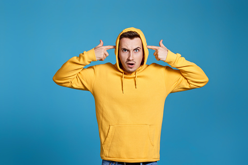 shocked man in yellow sweatshirt holding his fingers on his temples isolated on blue background