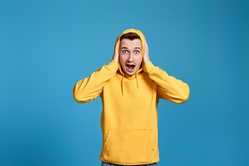 shocked man with round eyes, open mouth and hands isolated on blue background