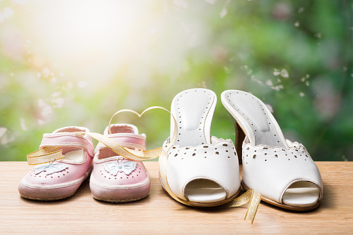 still life photography : mother and daughter shoes with heart shape gold ribbon on wood and blurred sunrise behind the bush background, love and bound concept