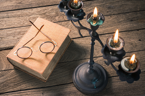 burning candles on handicraft brass candelabrum with vintage eyeglasses and old book on old wooden table, selected focus at the eyeglasses