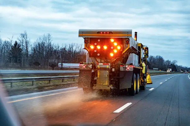 Car driver POV about to move past a state transportation department snowplow dump truck driving in the icy left lane on the New York State Thruway highway. The large commercial vehicle has a bright, flashing emergency light directional arrow pointing to the right on the back of the rig to warn faster following traffic to pass cautiously to the right while it spreads and scatters road salt with its automatic spreader equipment. Early January winter weather in western New York State, USA.