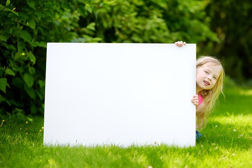 Cute little girl holding big blank whiteboard on warm and sunny summer day outdoors